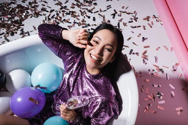 Vue de dessus de femme asiatique heureuse couchée dans la baignoire avec des ballons et tenant un verre de champagne près de confettis — Photo de stock