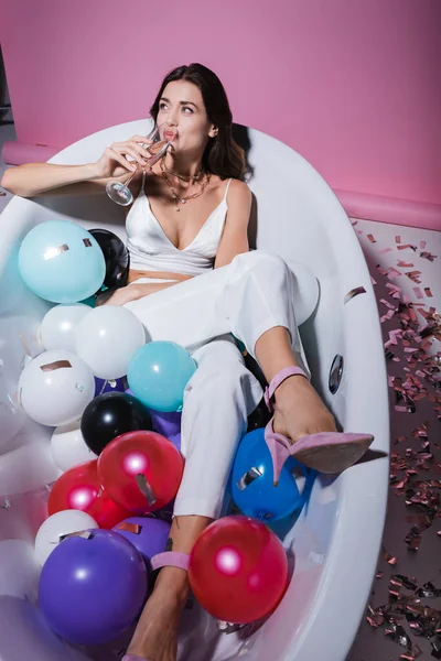 Щаслива жінка лежить у ванні з різнокольоровими кульками і п'є шампанське на рожевому — стокове фото