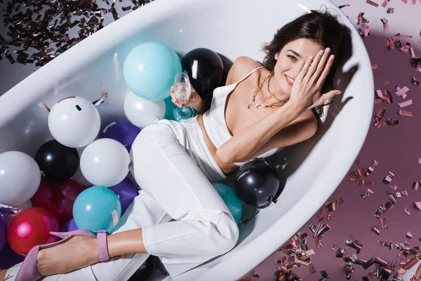 Vista superior de la mujer feliz cubriendo el ojo y acostado en la bañera con globos mientras sostiene la copa de champán - foto de stock