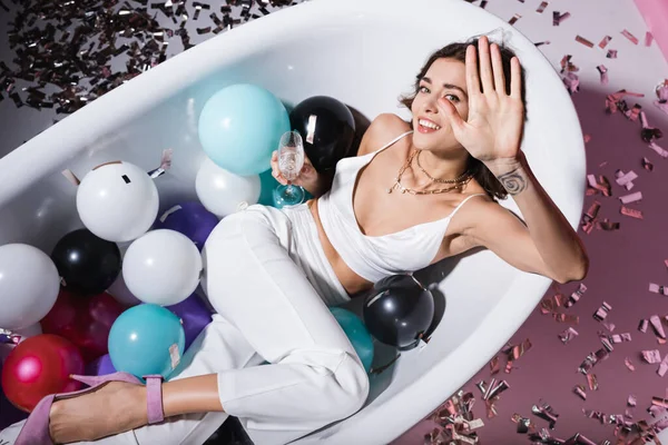Vue de dessus de la femme heureuse avec geste de tatouage et couché dans la baignoire avec des ballons tout en tenant un verre de champagne — Photo de stock