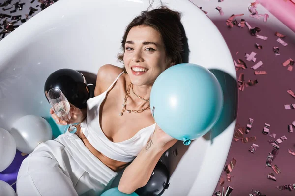 Top view of cheerful woman with tattoo gesturing and lying in bathtub with balloons while holding glass of champagne — Stock Photo