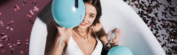 Вид зверху усміхненої жінки з татуюванням, що лежить у ванні, тримаючи повітряну кулю та келих шампанського, банер — стокове фото