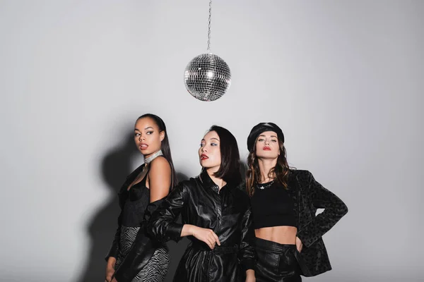 Young woman in leather beret posing with hand on hip near interracial models and shiny disco ball on grey — Stock Photo