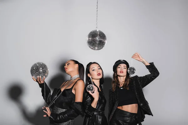 Young woman in leather beret posing near interracial models holding disco balls on grey — Stock Photo