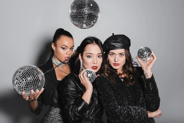 Young woman in leather beret posing near multiethnic models holding disco balls on grey — Stock Photo