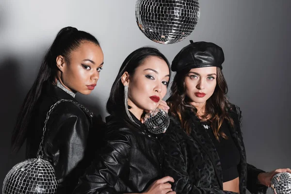 Young woman in beret looking at camera near interracial models holding disco balls on grey — Stock Photo