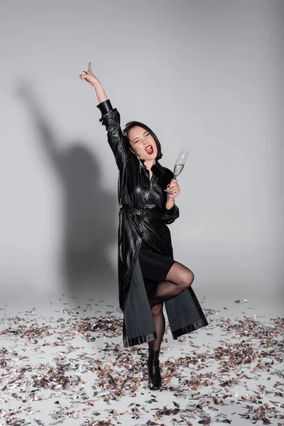 Full length of amazed asian woman holding glass of champagne while standing on shiny confetti on grey — Stock Photo