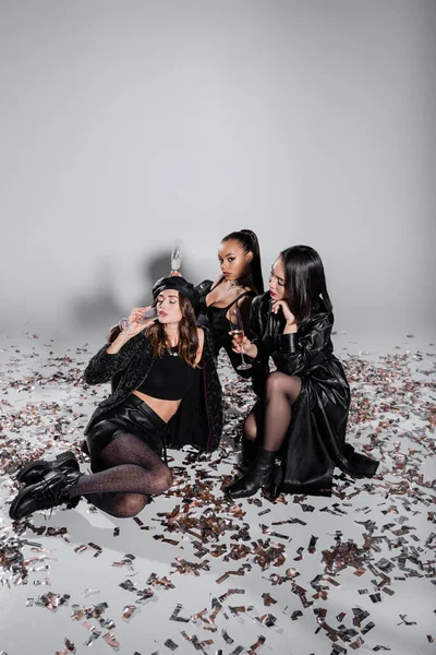 Longitud completa de jóvenes modelos multiétnicos sosteniendo copas de champán mientras se sienta cerca de confeti brillante en gris - foto de stock