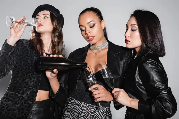 Young african american woman holding bottle and pouring champagne near multiethnic models with glasses on grey — Stock Photo