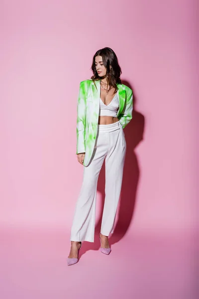 Full length of young brunette woman in blazer and white pants standing on pink — Stock Photo