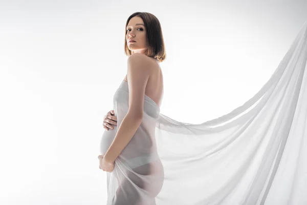 Morena mulher grávida em tecido tocando barriga isolada em cinza — Fotografia de Stock