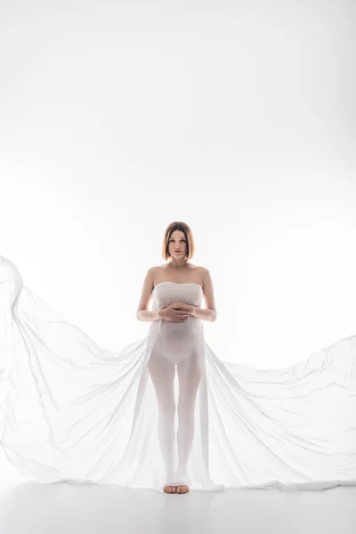 Young pregnant woman in white fabric looking at camera on grey background — Stock Photo