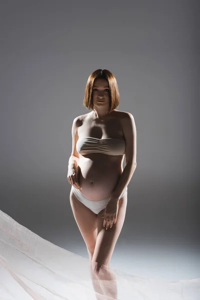 Donna incinta in biancheria intima posa vicino panno su sfondo grigio — Foto stock