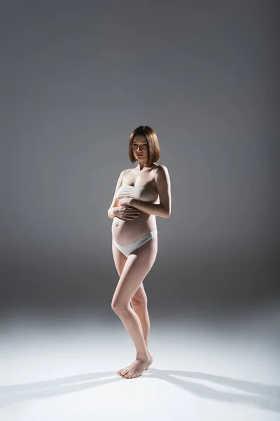 Босонога вагітна жінка в нижній білизні дивиться на камеру на сірому фоні — стокове фото