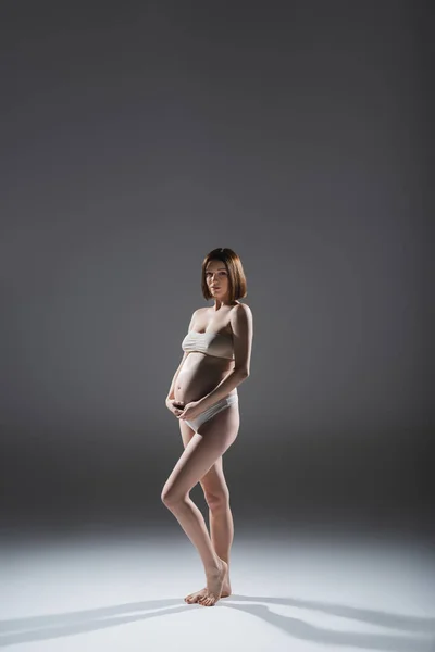 Giovane donna incinta in biancheria intima posa su sfondo grigio — Foto stock