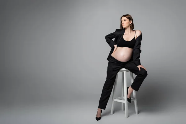 Pregnant woman in black suit posing on chair on grey background — Stock Photo