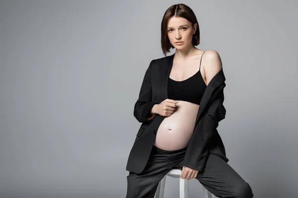 Stylish pregnant woman in top and suit sitting on chair isolated on grey — Stock Photo