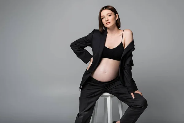 Jeune femme enceinte en costume noir et haut assis sur chaise isolé sur gris — Photo de stock