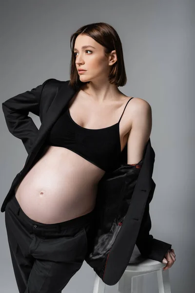 Stylish pregnant woman in suit looking away near chair isolated on grey — Stock Photo