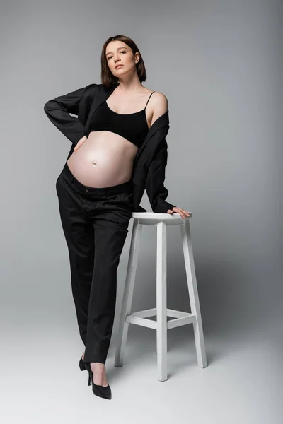 Jeune femme enceinte en costume et talons posant près de la chaise sur fond gris — Photo de stock