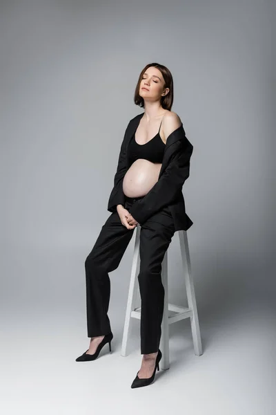 Embarazada modelo en traje cerrando los ojos mientras está sentado en la silla sobre fondo gris - foto de stock