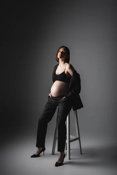 Stylish pregnant woman in heels and jacket sitting on chair on dark grey background — Stock Photo