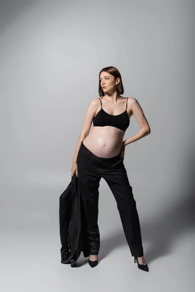 Fashionable pregnant woman in top holding jacket and posing on grey background — Stock Photo