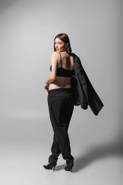 Молода модель зверху і підбори тримає куртку і дивиться на камеру на сірому фоні — стокове фото