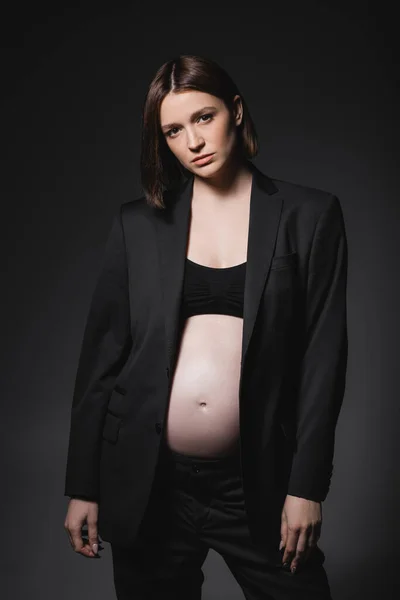 Brunette pregnant woman in suit looking at camera isolated on dark grey — Stock Photo