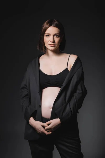 Young pregnant woman in black blazer looking at camera isolated on dark grey — Stock Photo