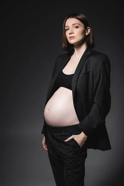 Young pregnant model in suit and top holding hand in pocket of pants isolated on dark grey — Stock Photo