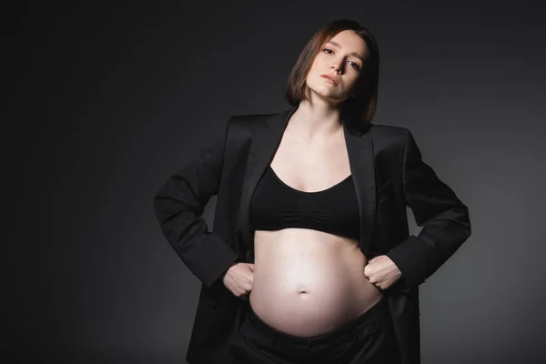Elegante donna incinta in giacca che si tiene per mano su fianchi isolati su grigio scuro — Foto stock