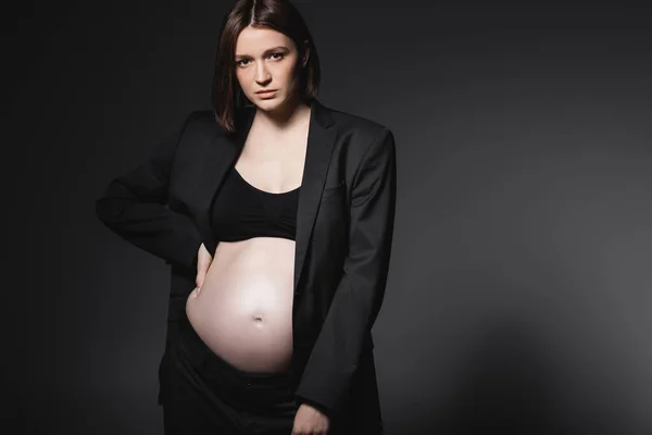 Mujer embarazada de moda en chaqueta negra cogida de la mano en la cadera aislada en gris oscuro - foto de stock