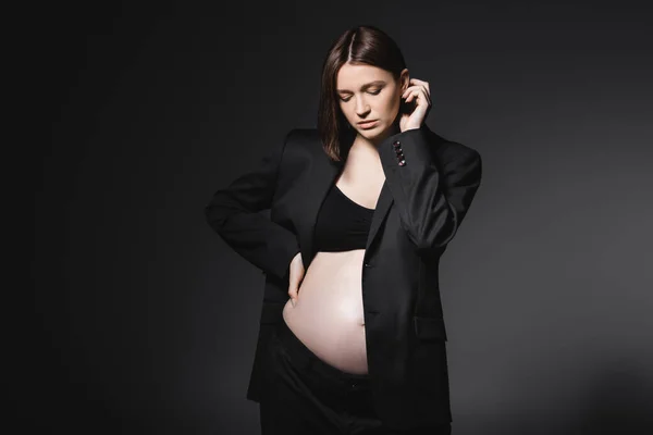 Mujer embarazada joven en la parte superior y chaqueta tocando el cabello aislado en gris oscuro - foto de stock