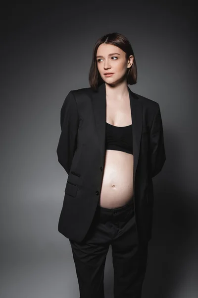 Mujer embarazada joven en top negro y chaqueta mirando hacia otro lado sobre fondo gris - foto de stock