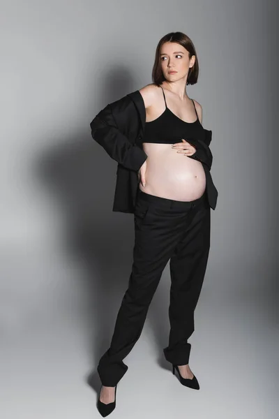 Full length of pregnant woman touching hip and belly on grey background — Stock Photo