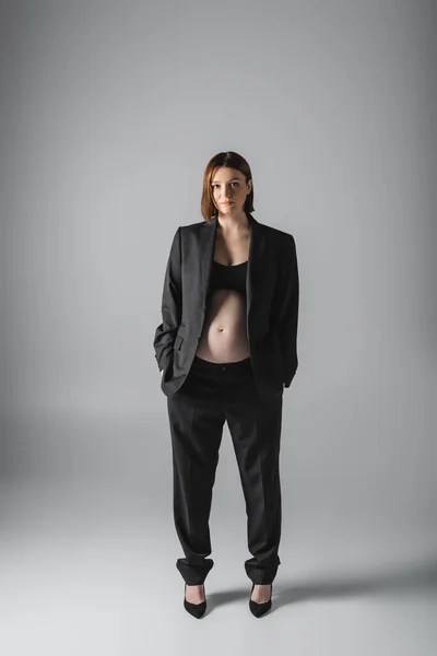 Young pregnant woman in suit and heels standing on grey background — Stock Photo