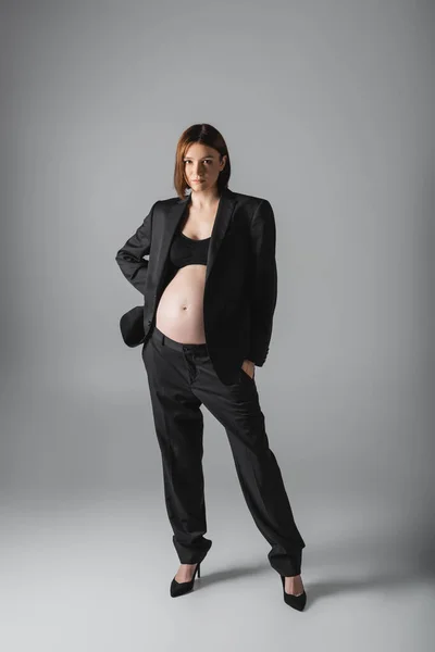 Молода вагітна жінка в костюмі і підбори дивиться на камеру, стоячи на сірому фоні — стокове фото
