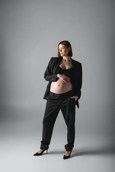 Mujer embarazada en traje y vientre superior tocando y sosteniendo la mano en el bolsillo sobre fondo gris - foto de stock