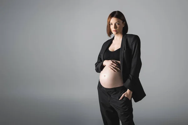 Pregnant woman in blazer and top touching belly isolated on grey — Stock Photo