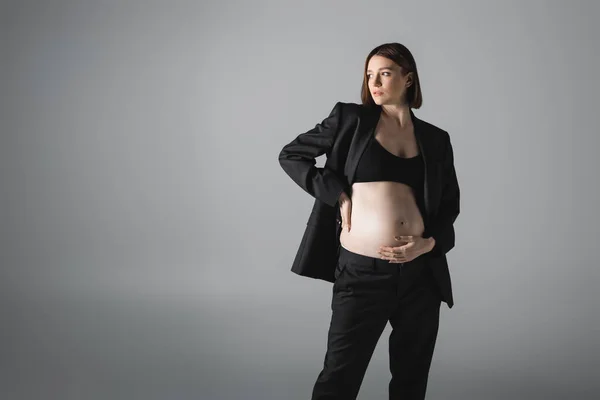 Стильная беременная женщина в топе и куртке, трогающей животик, изолированная от серого — стоковое фото