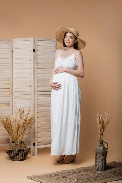Donna incinta sorridente in abito e cappello da sole in piedi vicino a schermo pieghevole sul beige — Foto stock