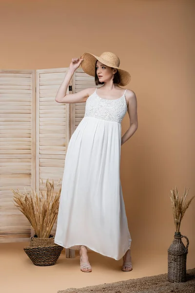 Stylish pregnant woman in dress and straw hat standing near plants and folding screen on beige — Stock Photo