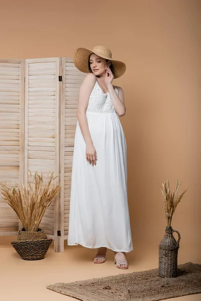Young pregnant woman in straw hat and dress standing near folding screen and spikelets on beige — Stock Photo