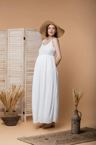 Donna incinta in abito e cappello da sole in piedi vicino a schermo pieghevole e spighette sul beige — Foto stock