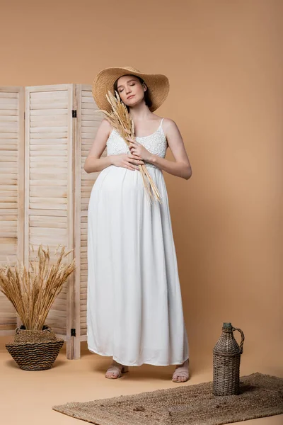 Stylish pregnant woman in dress and sun hat holding spikelets on beige — Stock Photo