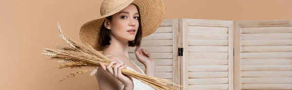 Mujer joven con sombrero de paja sosteniendo espiguillas cerca de la pantalla plegable aislada en beige, pancarta - foto de stock