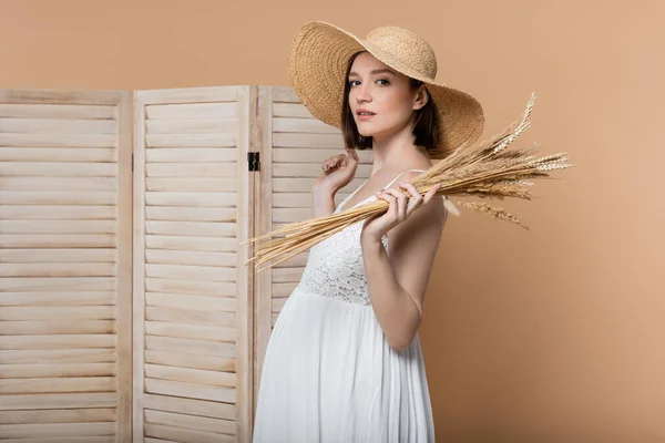 Mulher grávida em chapéu de sol segurando espiguetas e olhando para a câmera isolada no bege — Fotografia de Stock
