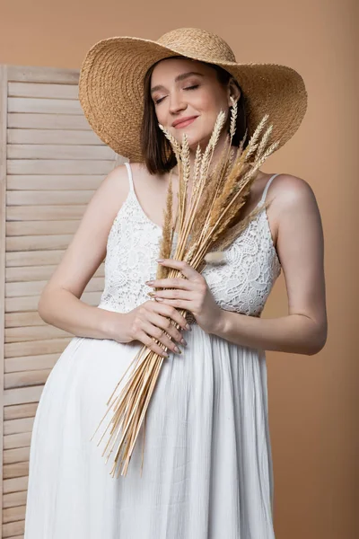 Smiling pregnant woman in dress holding spikelets near folding screen isolated on beige — Stock Photo