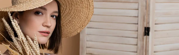 Portrait of woman in sun hat looking at camera near spikelets and folding screen on beige, banner — Stock Photo